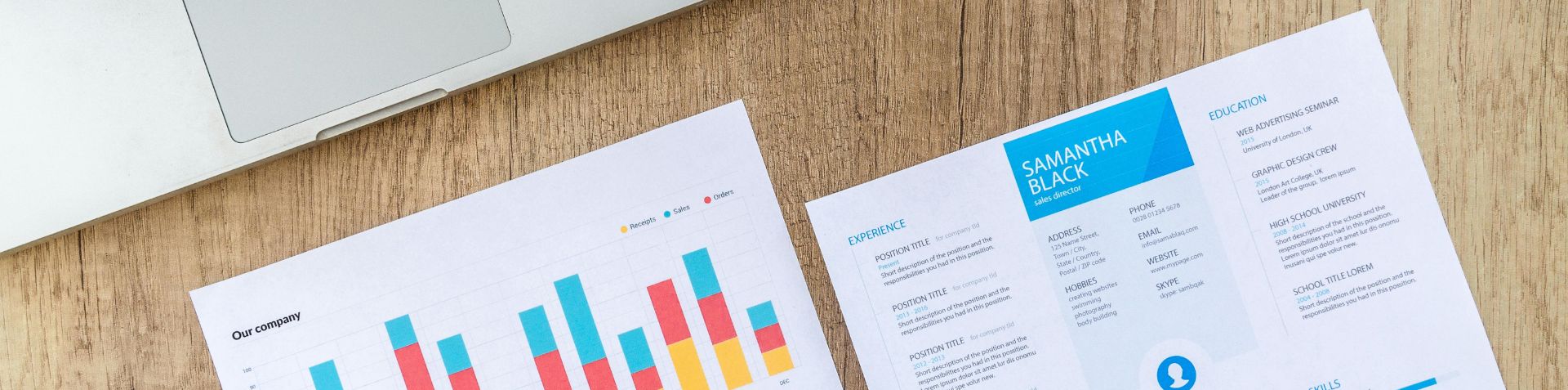 Two white sheets of paper, a CV and a business report, next to an open laptop on a wooden desk.